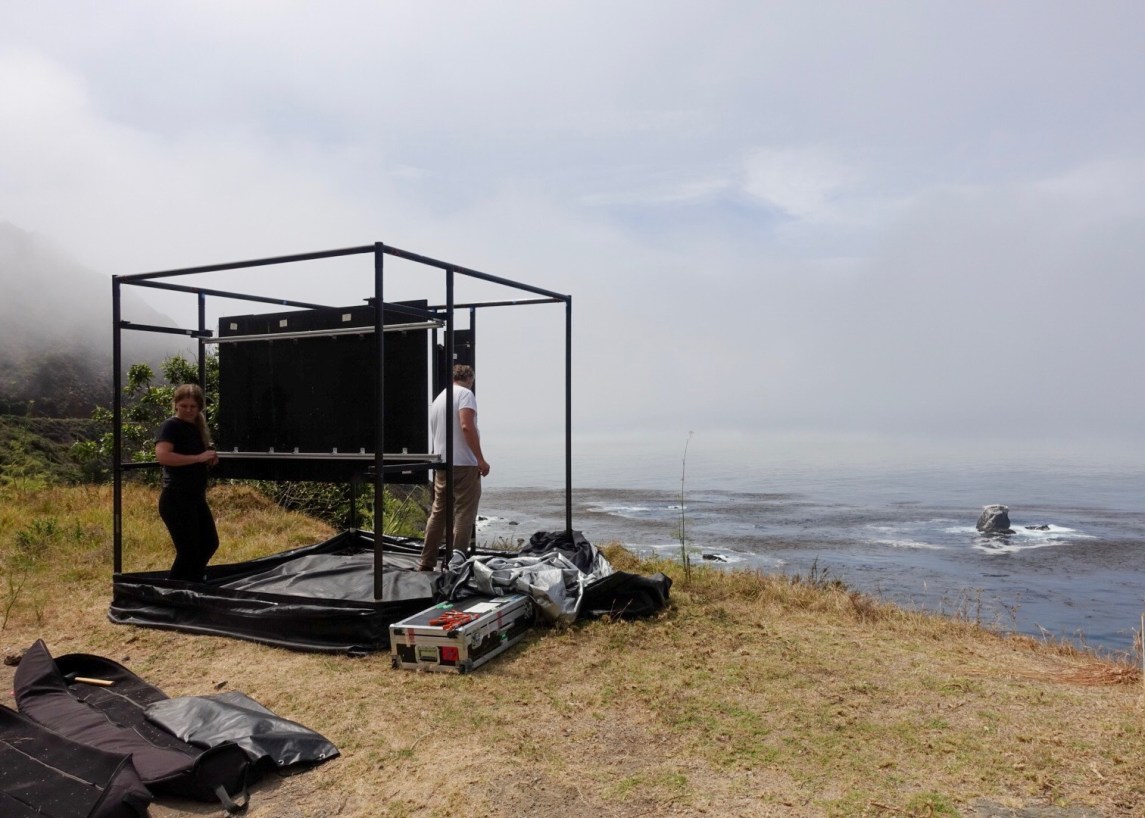 Color photograph of a large cube-shaped armature on the shoreline, with two persons inside.
