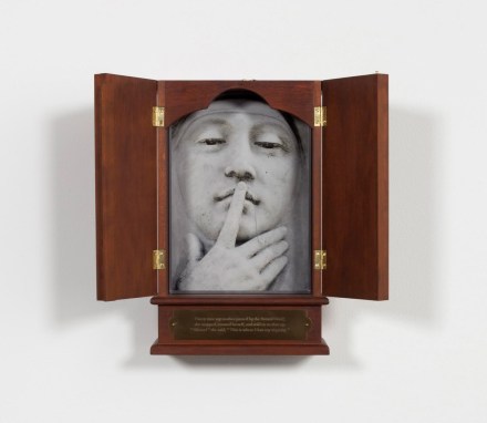 A wooden box with double doors, opened to reveal a photograph of a sculpture of a face with pointer finger to lips