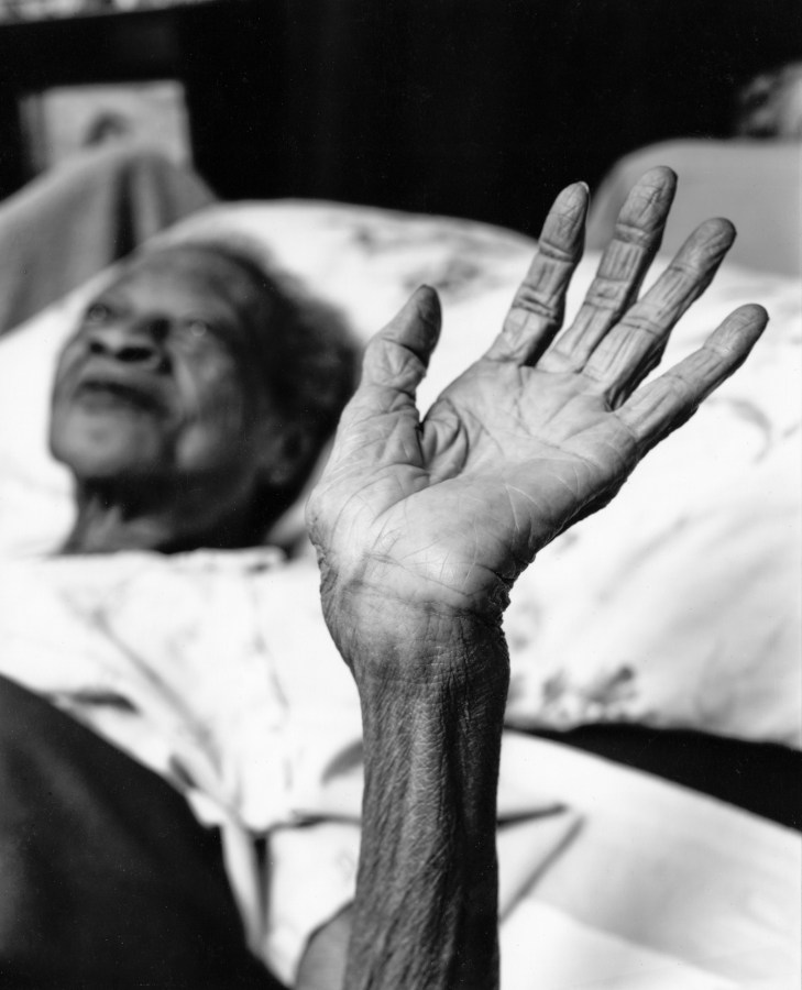 Black-and-white photograph of an elderly woman lying in bed raising her open hand to the viewew