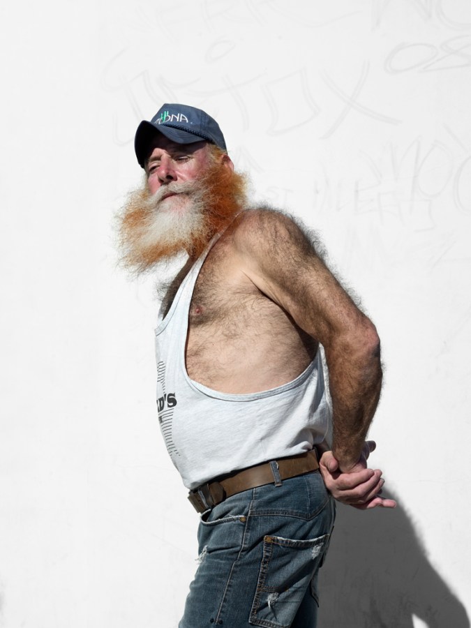 Color photograph of a man in a baseball cap and jeans standing with his hands clasped behind his back against a blank white wall