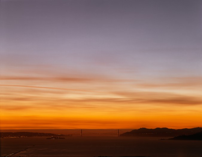 Color photograph of the Golden Gate Bridge on the horizon under a bright red-orange and violet sunset