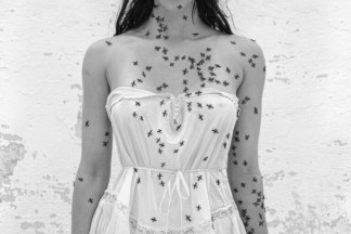 A black and white photograph shows a woman in a white negligee against a wall with chipped white paint. Small insects crawl on her body and on the flower-like object she holds in her hand.