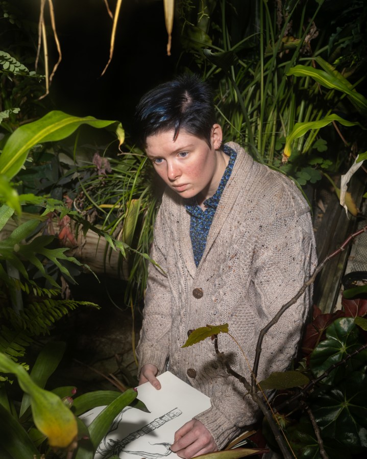 Color photograph of a figure surrounded by plants sketching on a sketch pad with charcoal