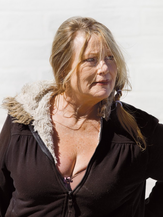 Color photographic portrait of a woman in a black jacket standing in front of a blank white wall