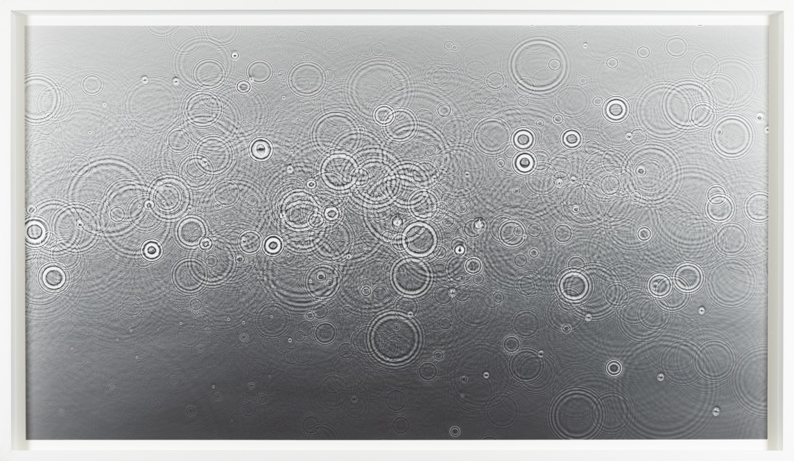 Color image of a grey toned photogram depicting water droplets cascading on the surface framed in white