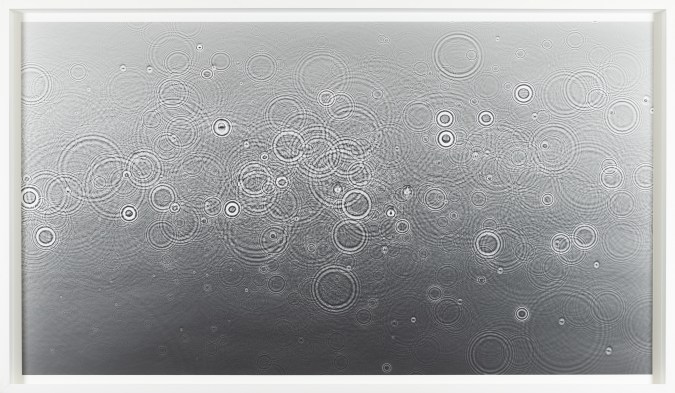 Color image of a grey toned photogram depicting water droplets cascading on the surface framed in white