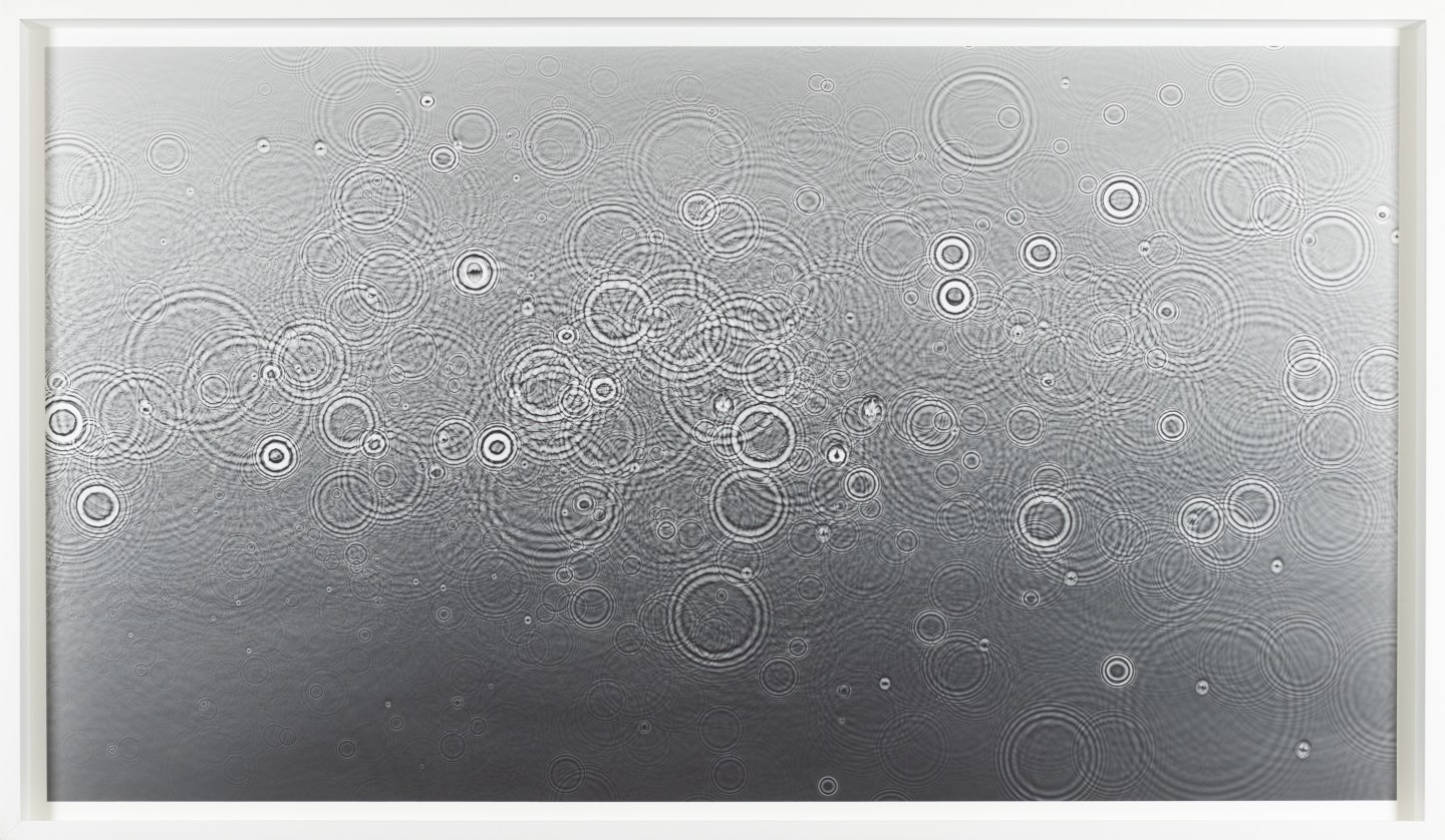 Color image of a grey toned photogram depicting water droplets cascading on the surface framed in white