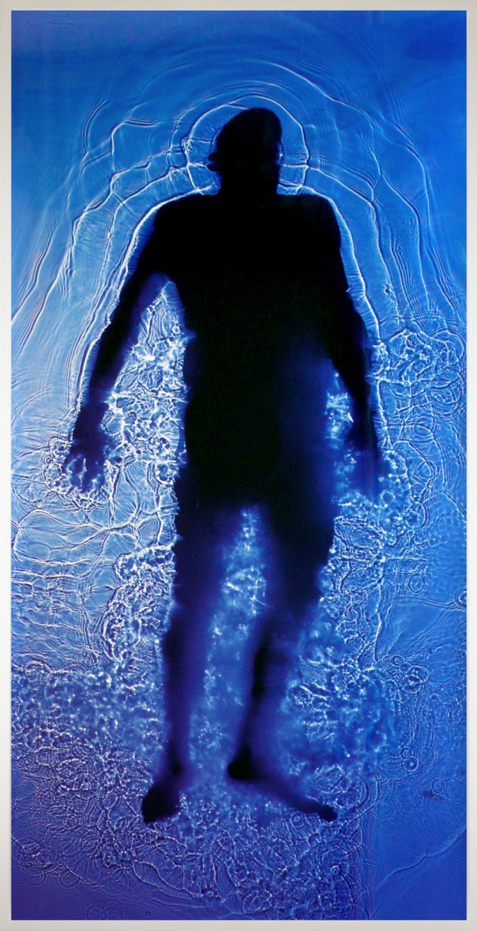 Color photograph of a man's full-body silhouette floating in a pool of transparent blue and violet liquid