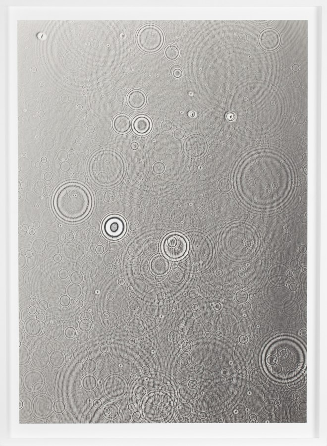 Color image of a grey toned photogram depicting water droplets cascading on the surface framed in white