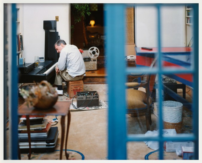 Color image of a man sitting on wooden box in front of piano through blue french doors in white frame