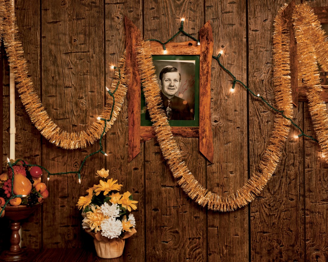 A color photograph of a wood-paneled room, with a black and white autographed portrait hung on the wall. Gold streamers are hung across it.