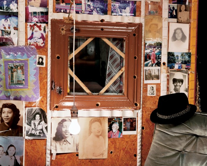 A color photograph of a framed mirror on a wall, surrounded by faded snapshots, with a lightbulb hanging from a wire in front of it.