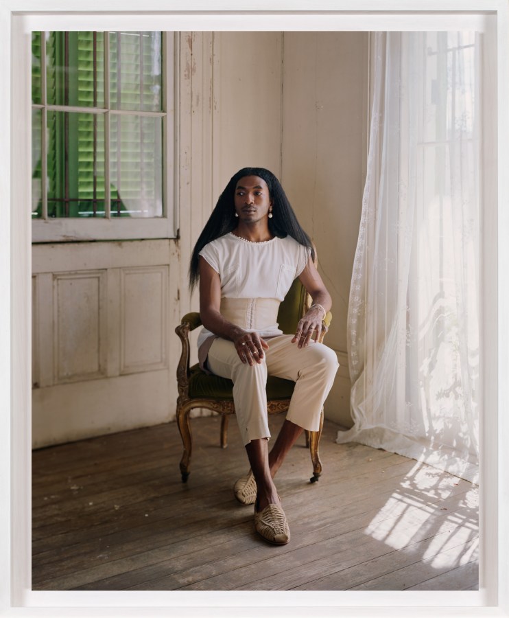A framed photograph of a person in a chair in the corner of a room with transparent white curtains