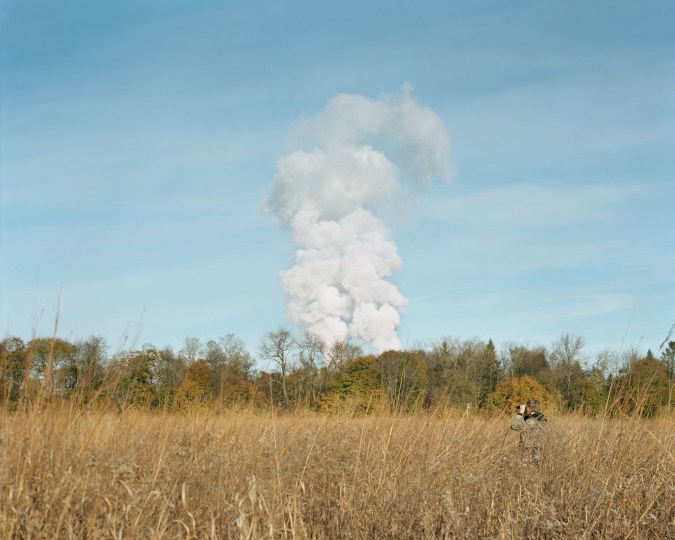Color image of a person with binoculars in grain field with large cloud of smoke on the horizon