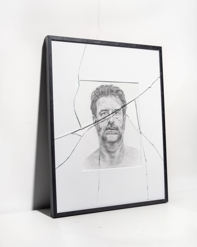 An image of a framed drawing leaning against a white wall depicts a man's face, the glass within the frame is severely cracked