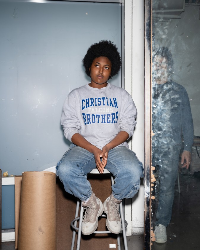 Color photograph of a seated figure with the reflection of the photographer present in a dirty mirror