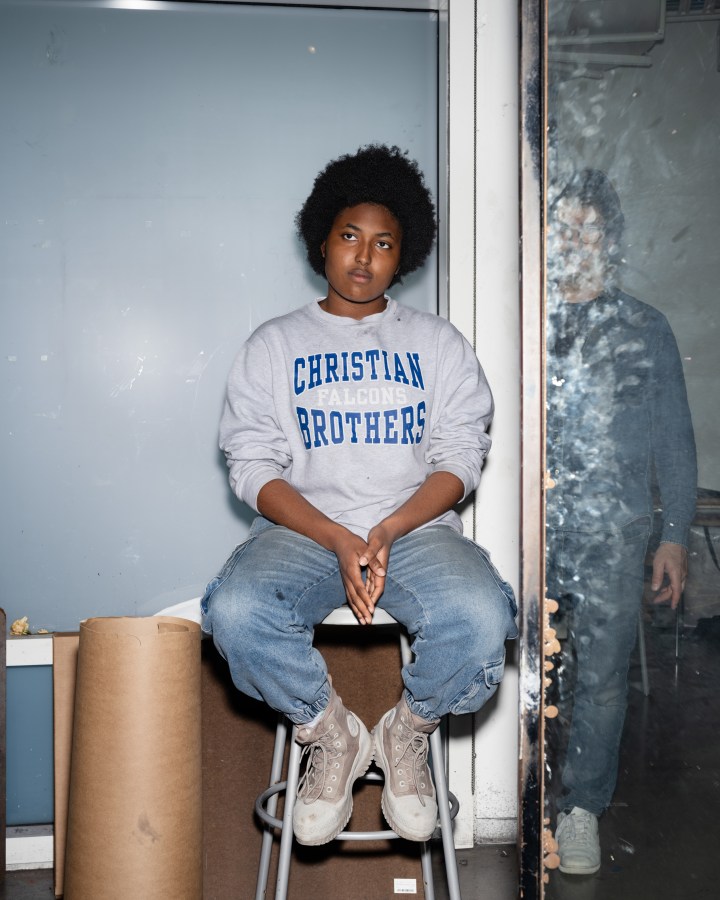 Color photograph of a seated figure with the reflection of the photographer present in a dirty mirror