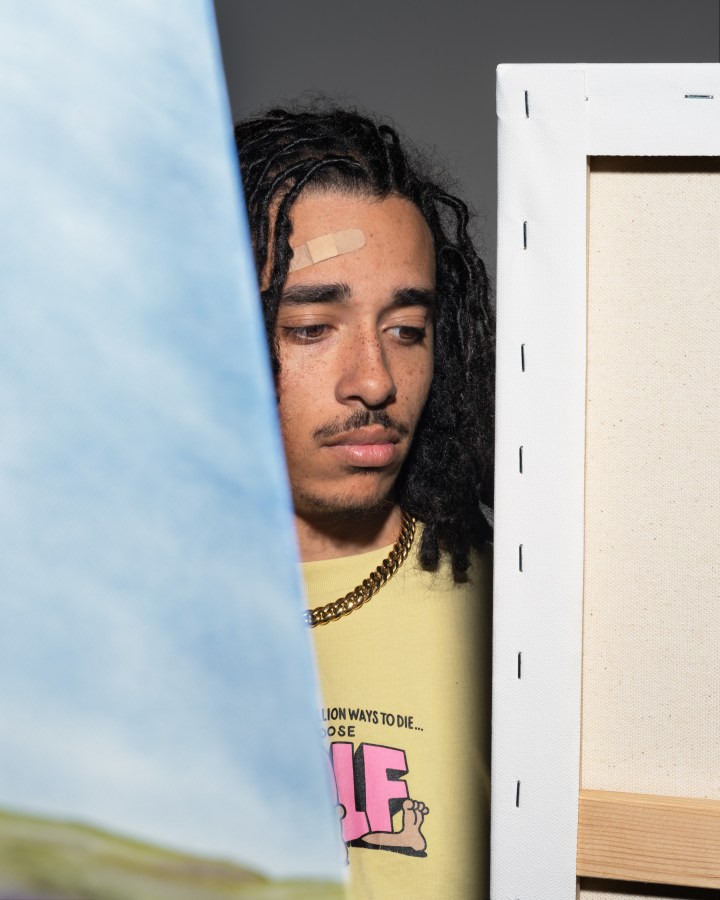 A color photograph shows a young man partially hidden behind two canvas paintings.
