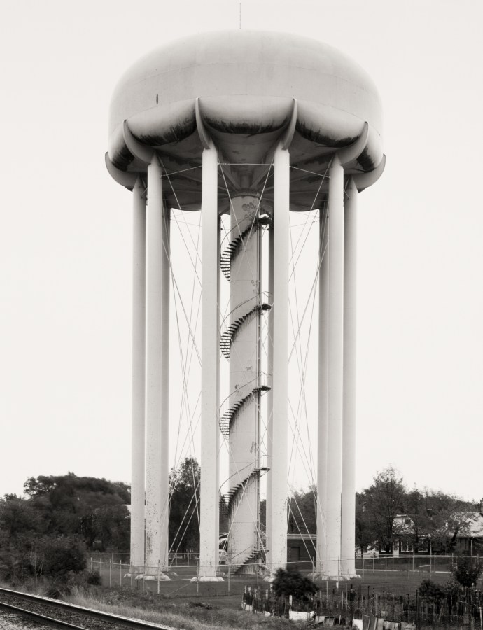 Black and white photograph of a water tower near a railroad track and residential homes