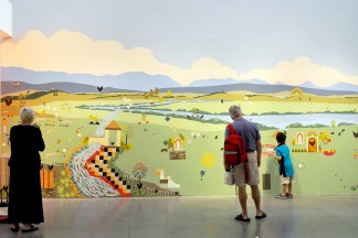 A color image of three people standing in front of a mural depicting an imaginary landscape