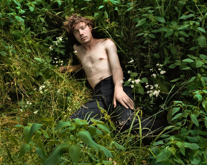 Color photograph of a shirtless man reclining in a grassy outdoor clearing
