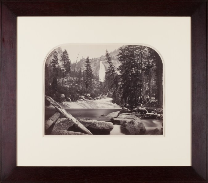 Sepia toned photograph of a waterfall centered amongst trees and rocks with lake in foreground framed in walnut