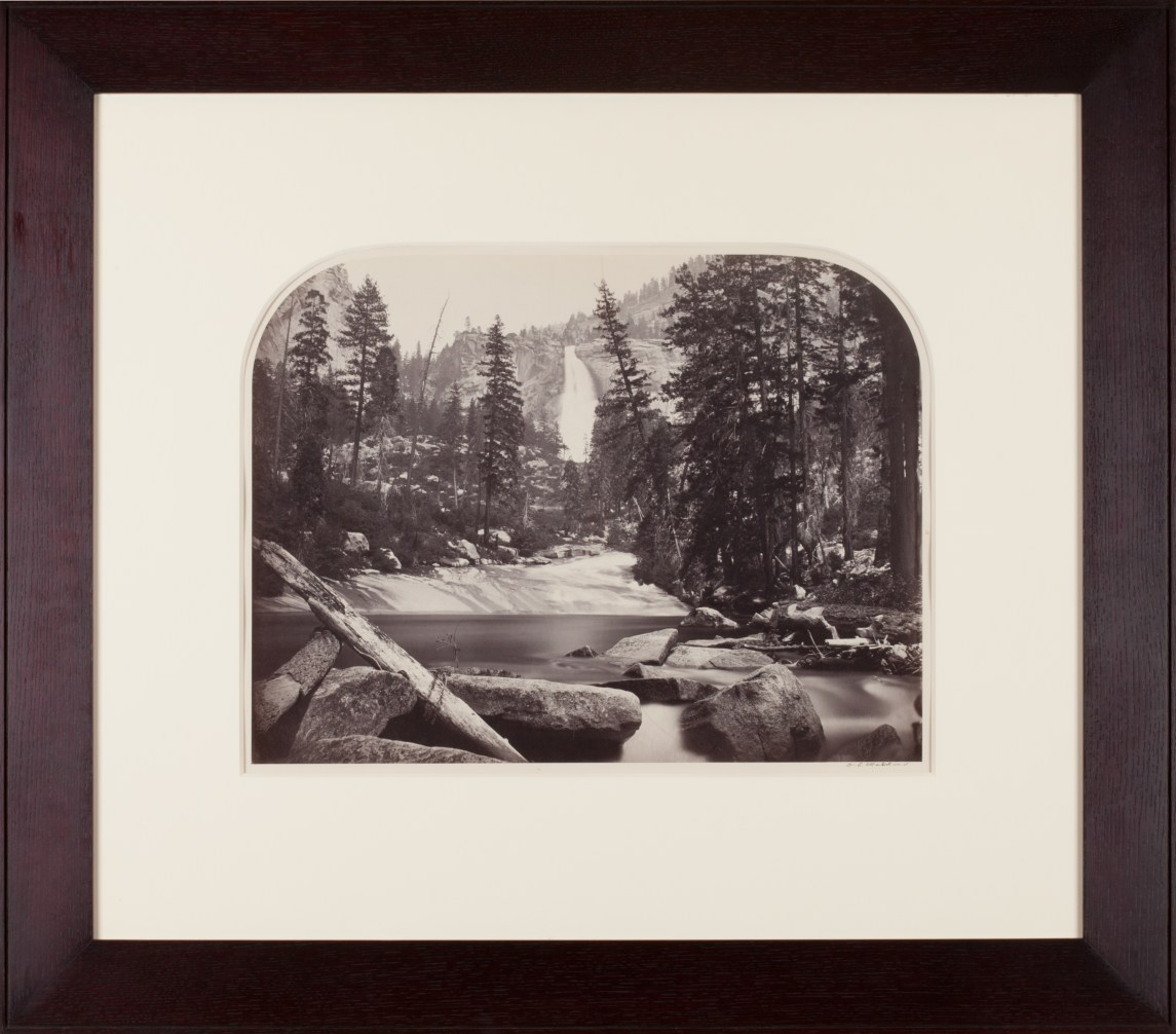 Sepia toned photograph of a waterfall centered amongst trees and rocks with lake in foreground framed in walnut