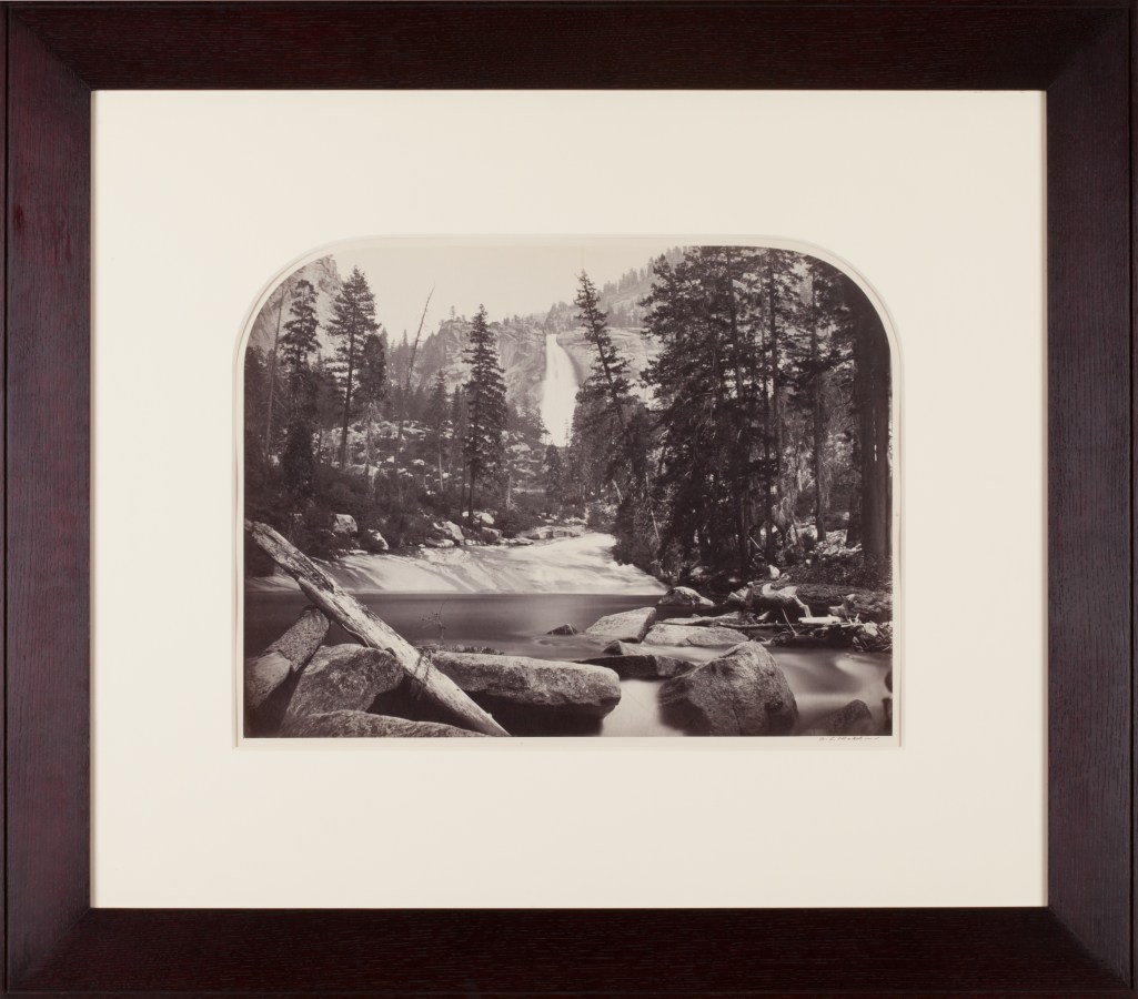 Sepia toned photograph of a waterfall centered amongst trees and rocks with lake in foreground framed in walnut