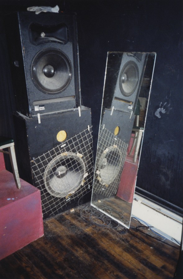 Color photograph of tower speakers next to a mirror leaning against a wall