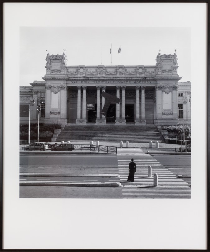 Image of a framed black and white photograph depicting a solitary black-clad woman walking through the crosswalk toward an imposing building.