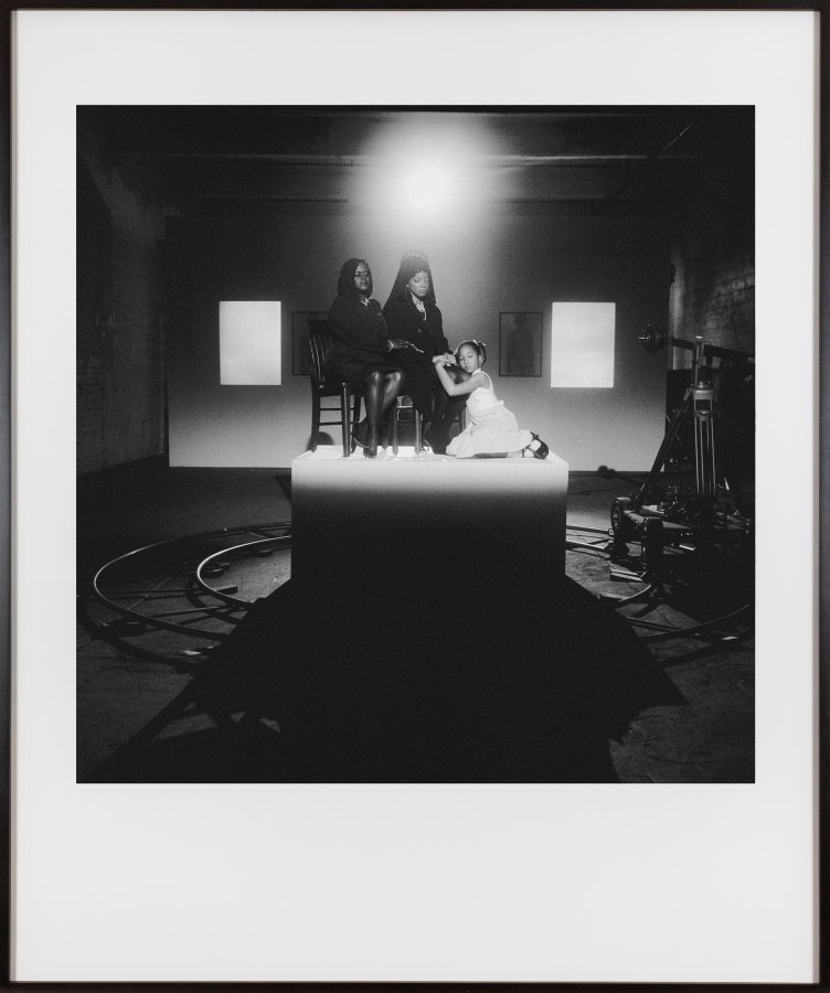 Framed black and white photograph of three people seated on a pedestal lit by a spotlight in a darkened room