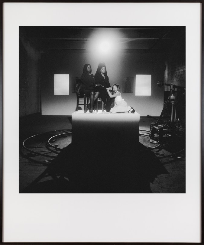 Framed black and white photograph of three people seated on a pedestal lit by a spotlight in a darkened room