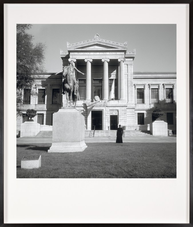 Black and white photograph of a figure in a black dress facing the exterior of a museum framed in black