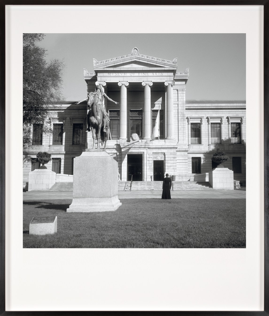 Black and white photograph of a figure in a black dress facing the exterior of a museum framed in black