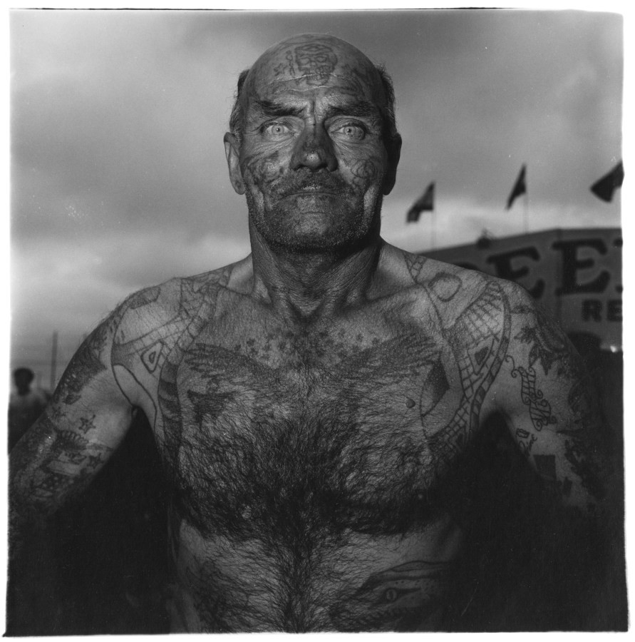 Black and white photograph of a shirtless tattooed man with flags and storm clouds in the background