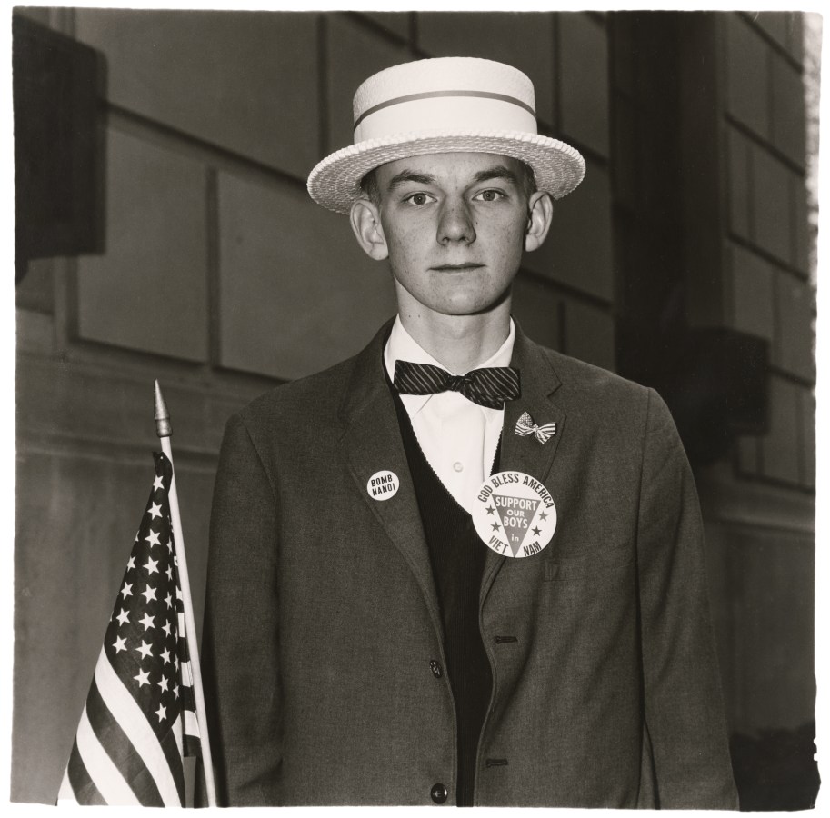 A black and white image features a young man wearing a straw hat and patriotic pins on his blazer while holding an American flag