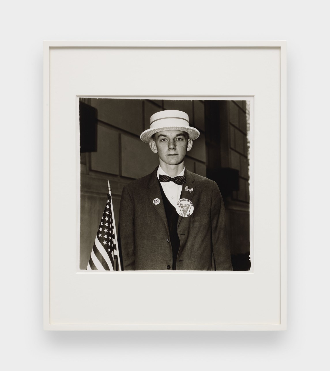 A black and white image features a framed photograph hanging on a white wall. The image depicts a young man wearing a straw hat and patriotic pins on his blazer while holding an American flag