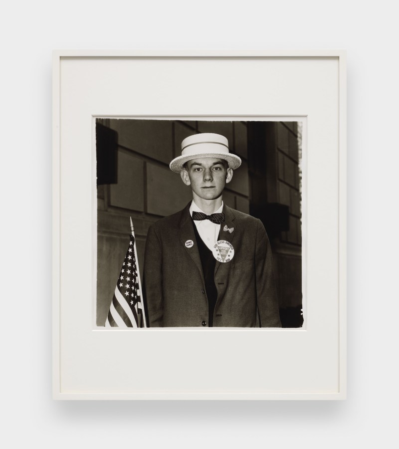 A black and white image features a framed photograph hanging on a white wall. The image depicts a young man wearing a straw hat and patriotic pins on his blazer while holding an American flag