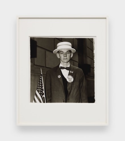 A black and white image features a framed photograph hanging on a white wall. The image depicts a young man wearing a straw hat and patriotic pins on his blazer while holding an American flag