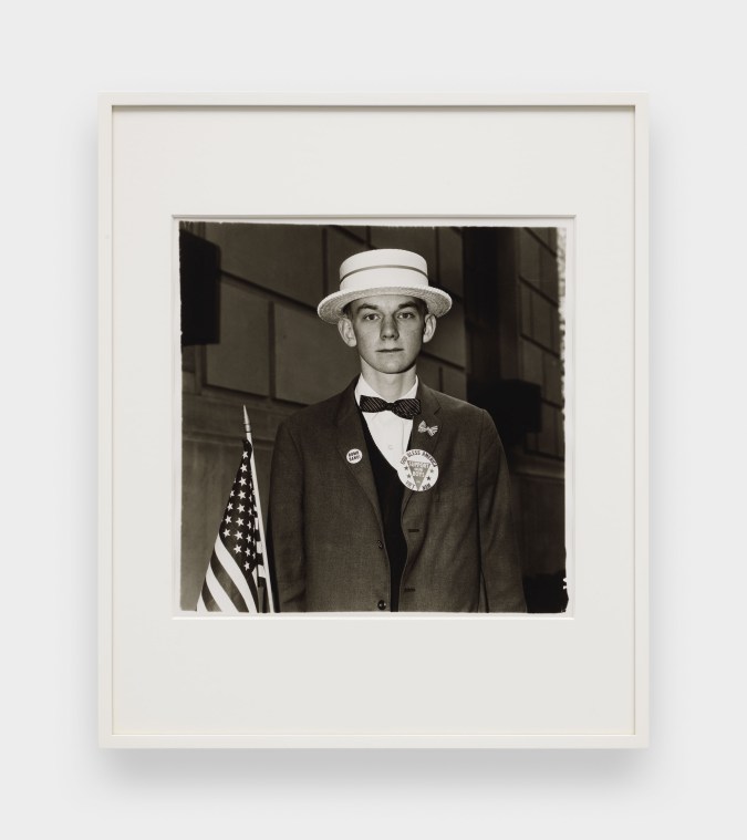 A black and white image features a framed photograph hanging on a white wall. The image depicts a young man wearing a straw hat and patriotic pins on his blazer while holding an American flag