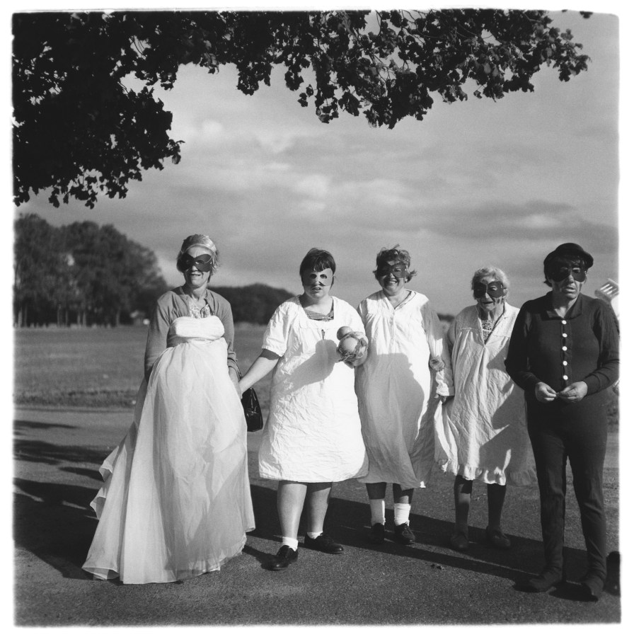 Black-and-white photograph of five figures wearing masks with an open field in the background and cloudy sky