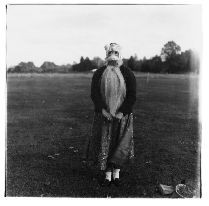 Black-and-white photograph of a figure wearing a mask with a field in the background