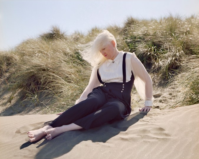 Color photograph of a woman with long white hair blowing in the wind seated on a sand dune covered with beach grasses