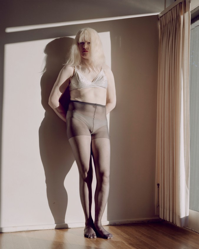 Color photograph of a woman with long white hair leaning against a wall next to a curtained window
