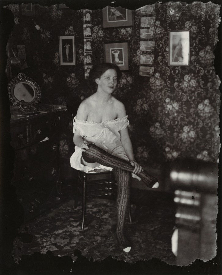 Black-and-white photograph of a woman with her legs crossed sitting in a wallpapered room
