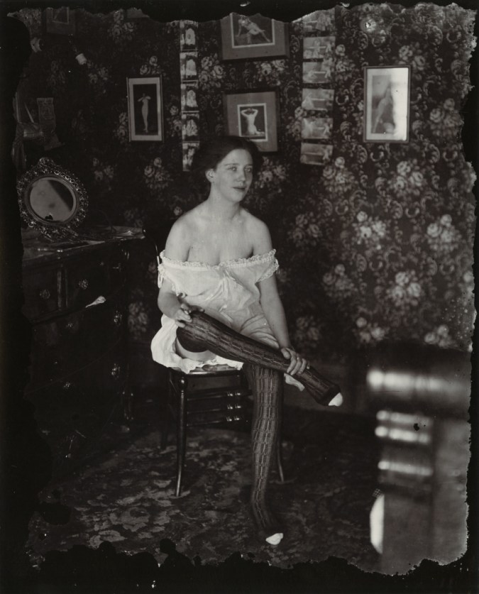 Black-and-white photograph of a woman with her legs crossed sitting in a wallpapered room