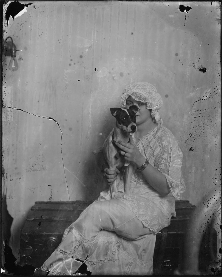 Black and white photograph of a woman in a white lace dress and white bonnet holding a small dog in her lap