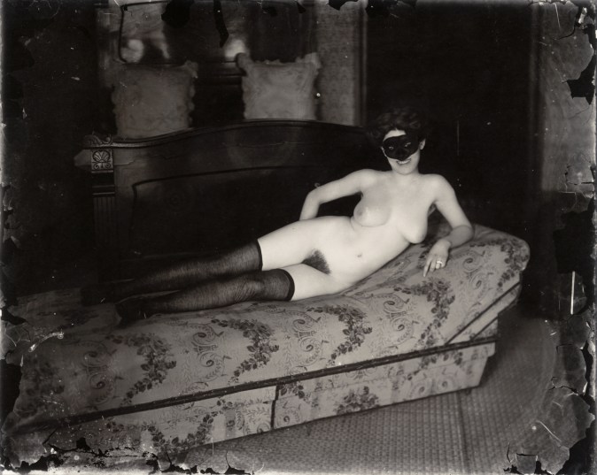 Black-and-white photograph of a nude woman wearing a mask laying on a sofa