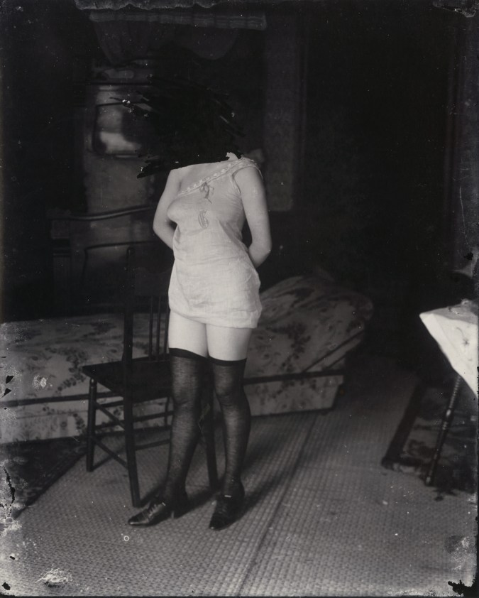 Black and white photograph of a woman in lingerie standing in front of a chair, with her face scratched out