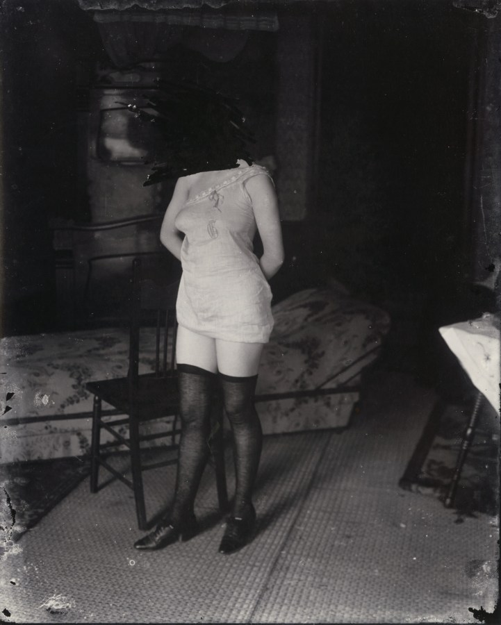 Black and white photograph of a woman in lingerie standing in front of a chair, with her face scratched out
